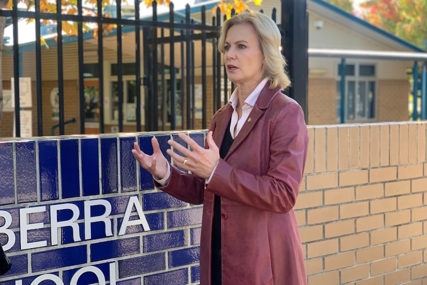 A blonde woman in a long leather coat stands outside a school, gesturing as she speaks.