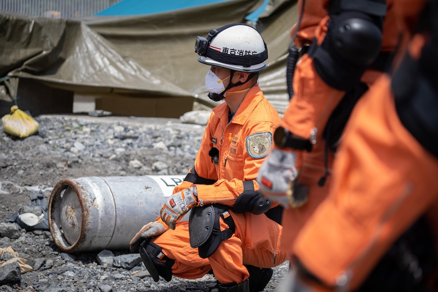 A man in orange overalls and white helmet kneels next to rubble. A patch on his arm shows a cartoon dog