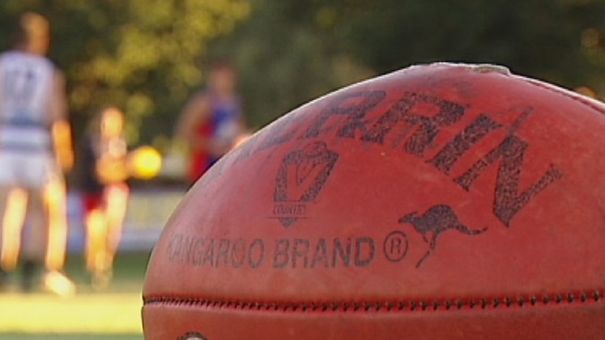 A close up of a red Sherrin brand Aussie Rules football, with blurred players in the background.