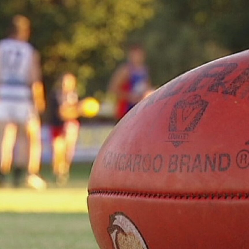 A close up of a red Sherrin brand Aussie Rules football, with blurred players in the background.