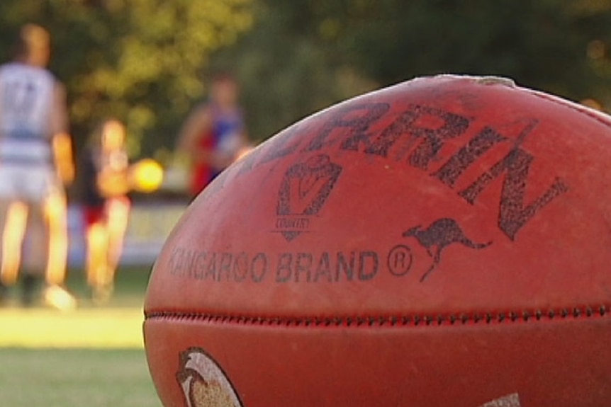 A close up of a red Sherrin brand Aussie Rules football, with blurred players in the background.