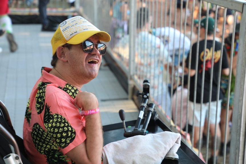 A young man wearing a pine shirt with pineapples and yellow cap is at a music festival. He uses a wheelchair.