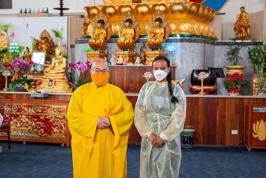 A bald man in yellow robes and a yellow mask stands with a woman in ppe gear and mask in front of gold Buddhist deities. 