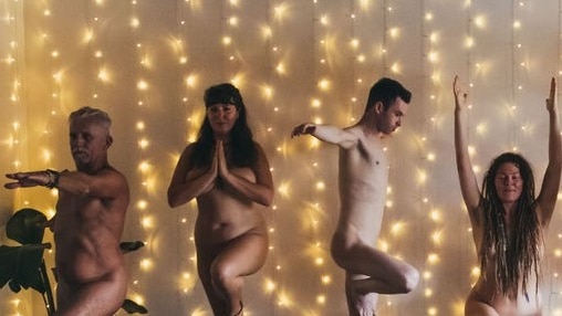 A group of naked people stand in yoga poses in front of a wall covered in fairy lights 