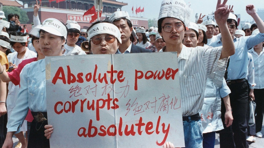 A group of Chinese journalists hold a sign that says "Absolute power corrupts absolutely" and wear hats made out of newspapers.