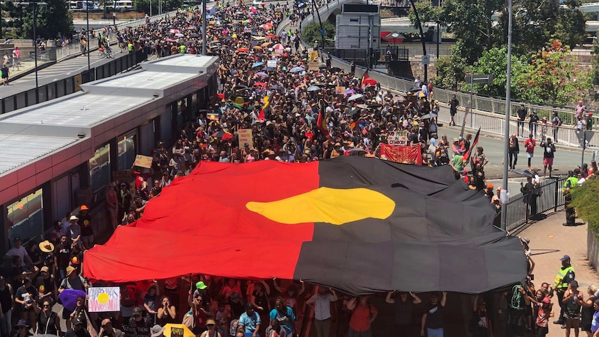 A giant Aboriginal flag is carried by members of a large crowd as they walk across a bridge as part of a protest.