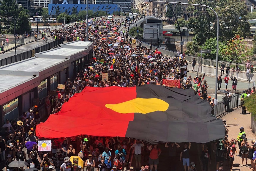 A giant Aboriginal flag is carried by members of a large crowd as they walk across a bridge as part of a protest.