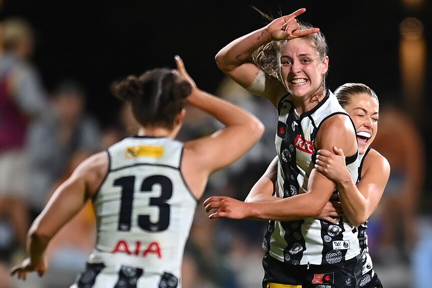 A Collingwood AFLW player celebrates a goal with two teammates.