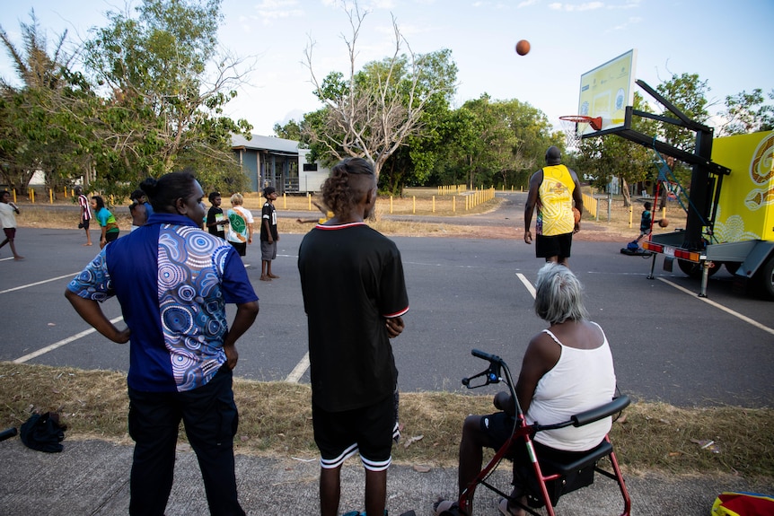Several adults watch on as young children shoot hoops at a basketball court.