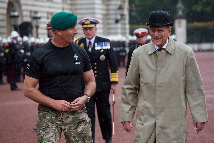 A man in a black t-shirt and green beret walks with Prince Philip, who wears a bowler hat and trench coat, in the rain.