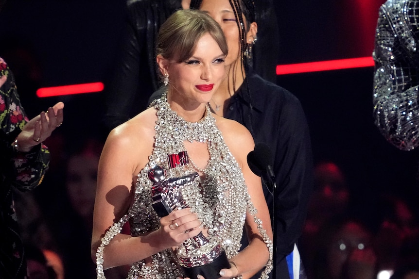 taylor swift smiles on stage standing behind a microphone holding a moonperson vma trophy