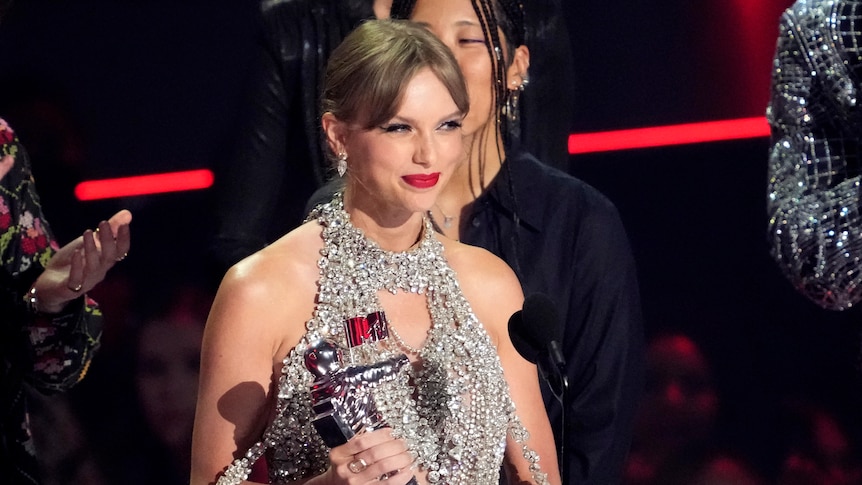 taylor swift smiles on stage standing behind a microphone holding a moonperson vma trophy