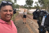 Gordon standing next to camera with Harriet in background holding microphone standing in front of swollen river.