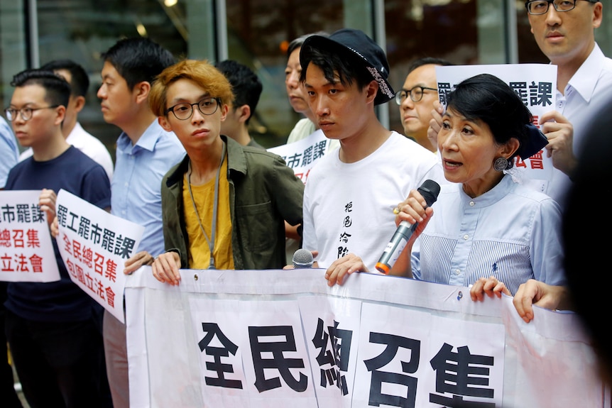 You see a group of Hongkongers protesting out of a glass-walled office behind banners with Chinese characters.