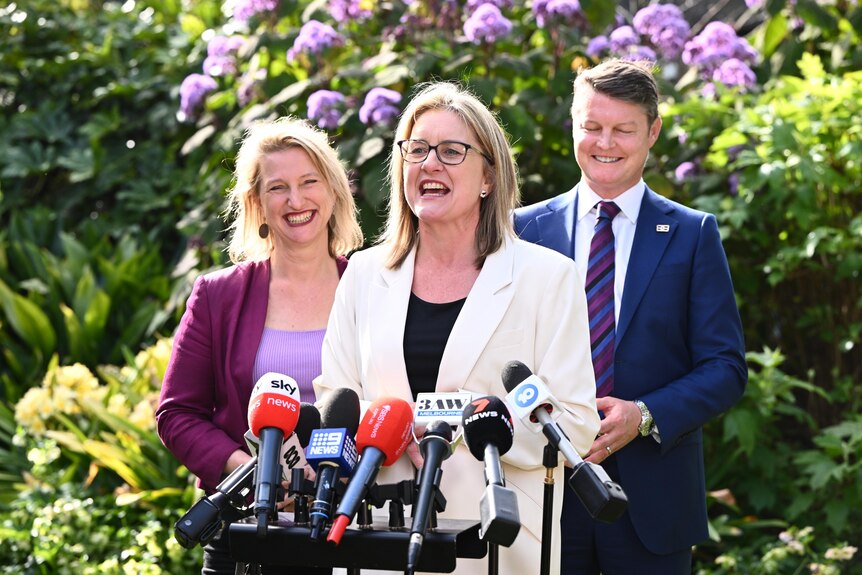 Two women and a man, all smiling, stand behind a bank of microphones.