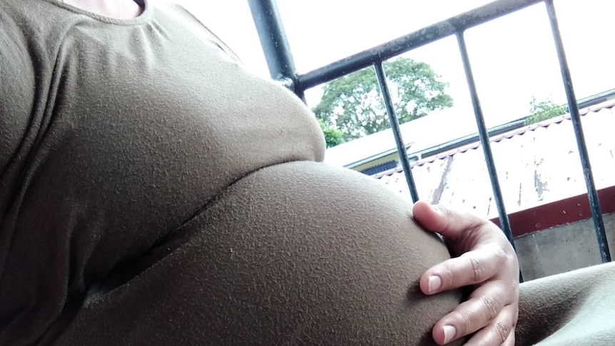 Heavily pregnant woman in brown dress, face not shown, hand on belly.