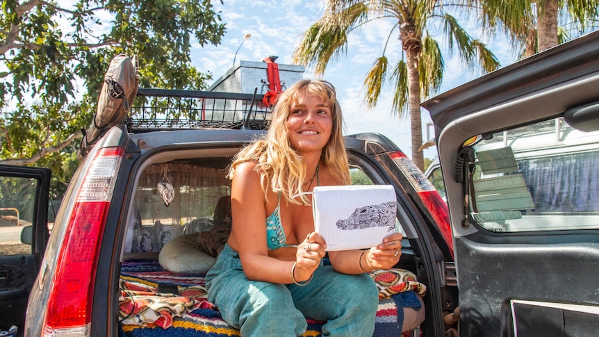 A young woman sits in the back of a camper vehicle holding her drawing