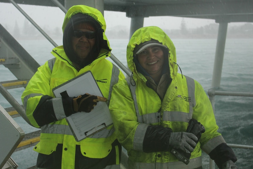 Two women wear raincoats and stand at the end of a jetty in the rain
