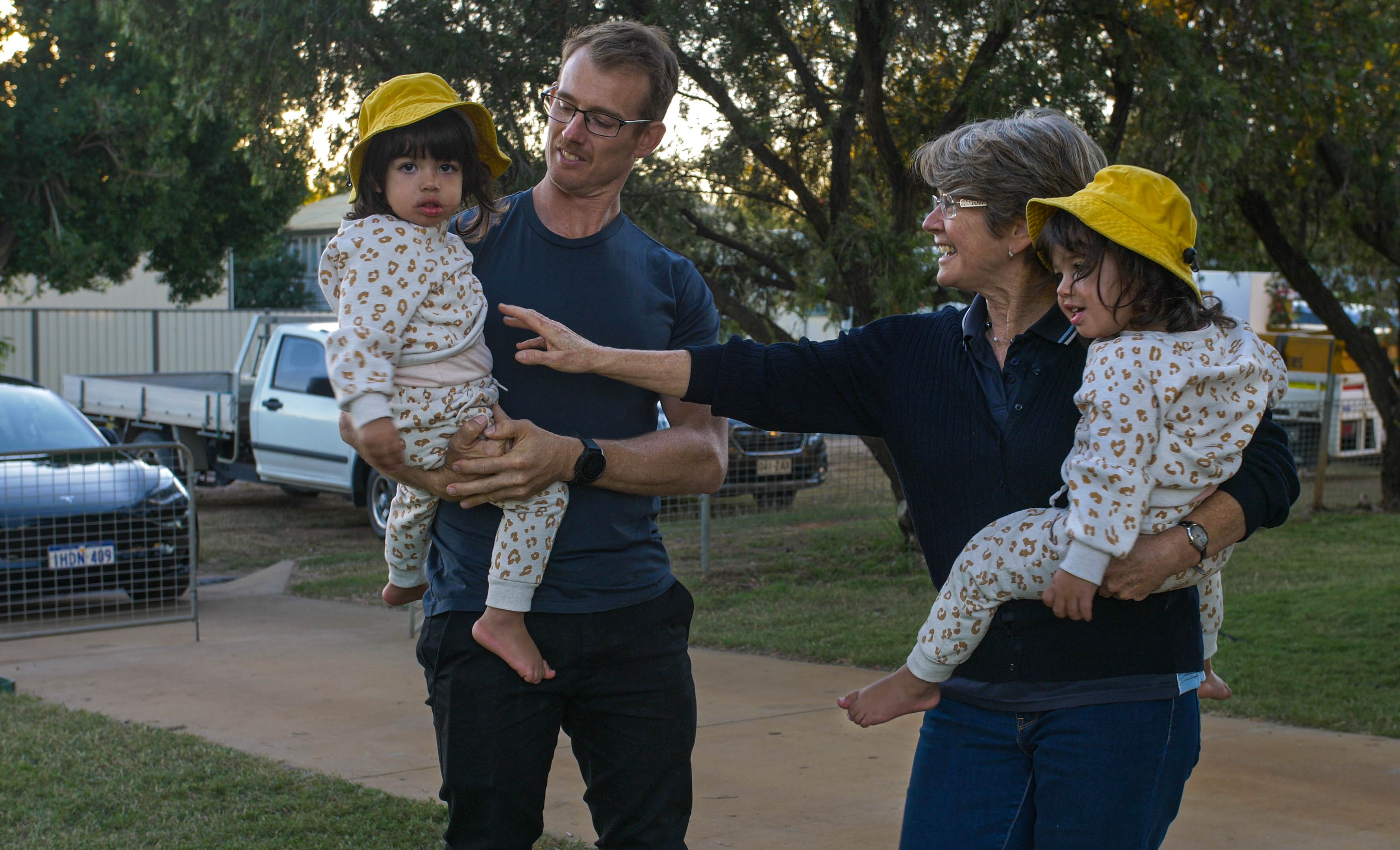 A man and a woman both hold an identical twin in their arms, the twins wearing matching outfits.