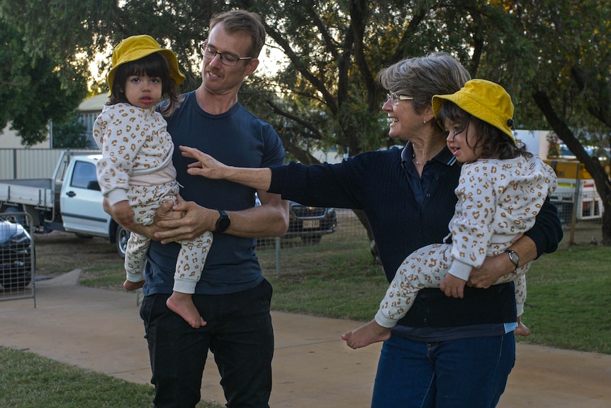 A man and a woman both hold an identical twin in their arms, the twins wearing matching outfits.