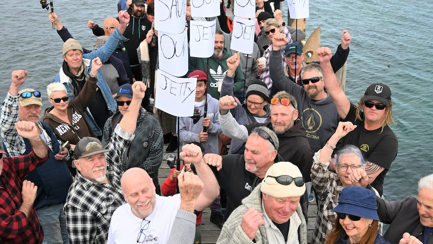 A group of people with raised fists and protest signs stand on a jetty 