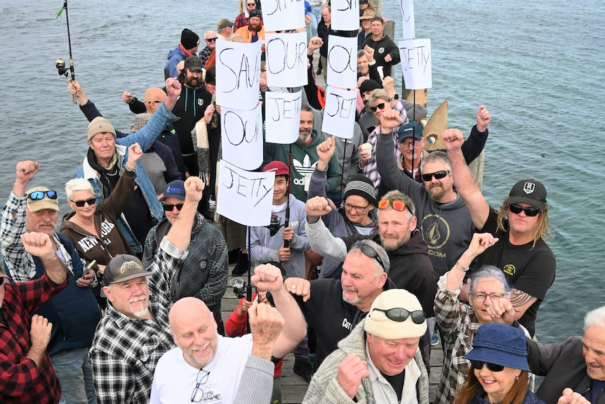 A group of people with raised fists and protest signs stand on a jetty 