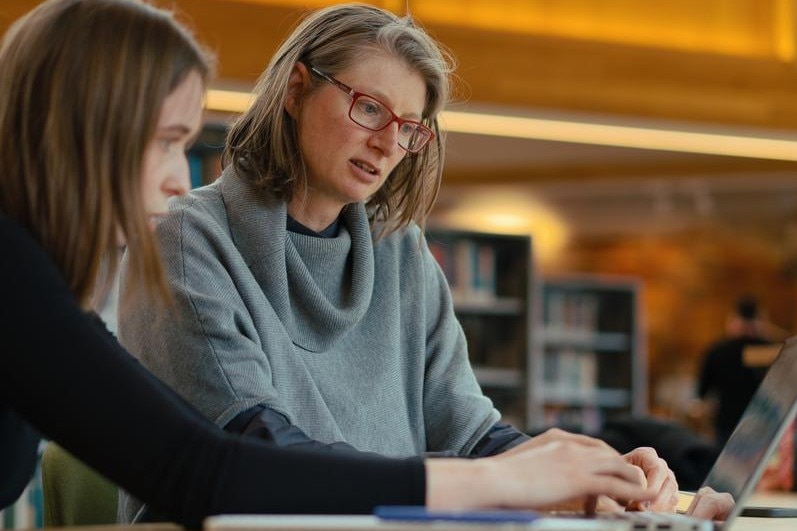 A woman with red glasses discusses something on a computer with a younger woman