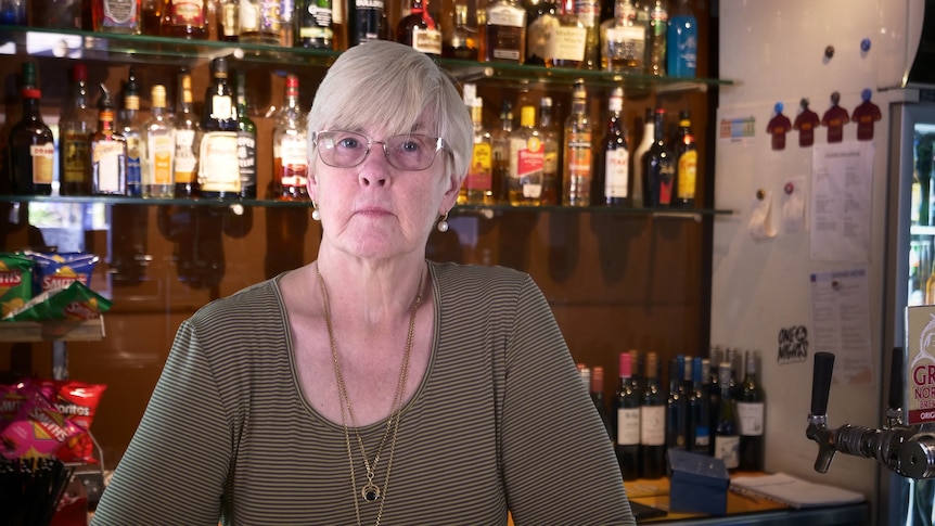 Diane Loechel stands behind a bar with lots of alcohol bottles. She has a serious expression.