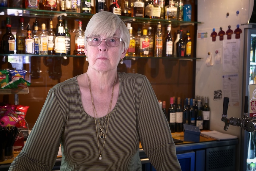 Diane Loechel stands behind a bar with lots of alcohol bottles. She has a serious expression.
