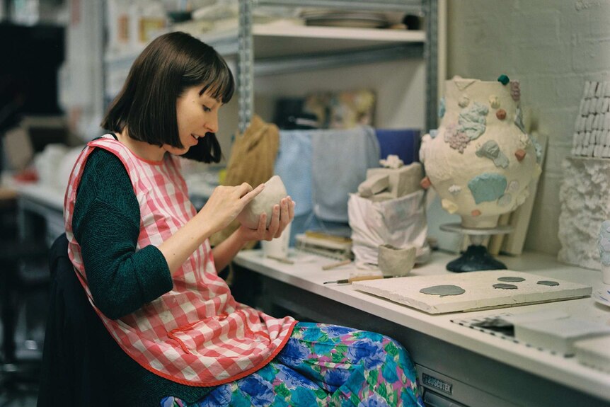 A woman with short dark hair sits at a desk moulding a clay bowl.