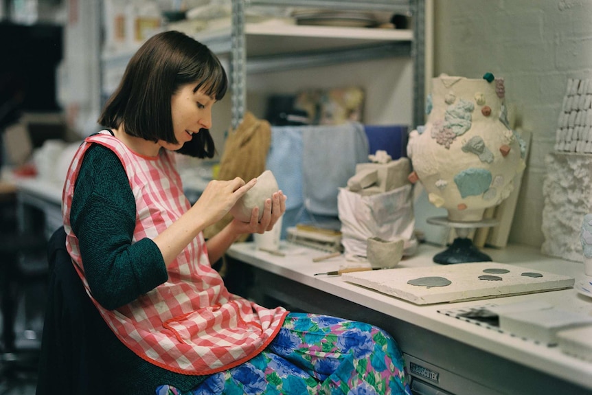 A woman with short dark hair sits at a desk moulding a clay bowl.