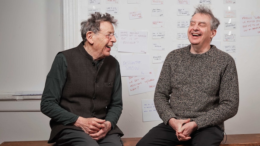 Philip Glass and Phelim McDermott sit on a bench laughing in front of a whiteboard.