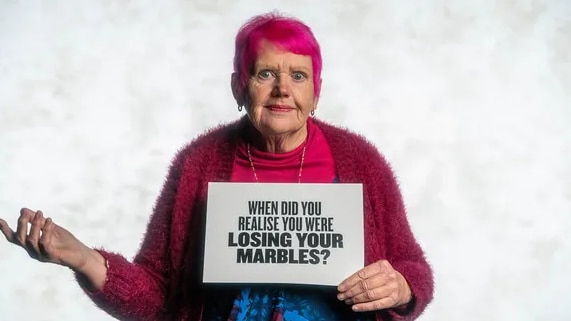 Older woman with pink hear and matching cardigan holds a sign that says "When did you realise you were losing your marbles?"