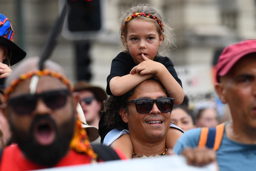 A little girl wearing a headband with colours of the Aboriginal flag sits on a man's shoulders at a rally