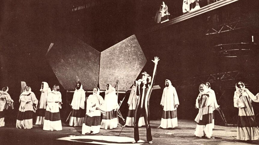 Wide photo of the stage and actors, with Jon English, arms raised, singing.