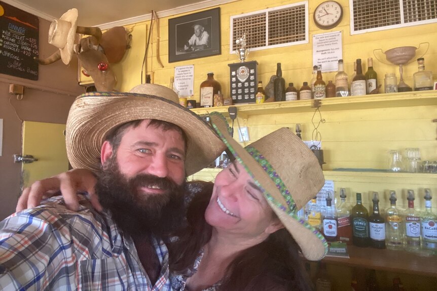 A man and a woman in cowboy hats smile for a selfie in front of a bar stacked with alcohol.