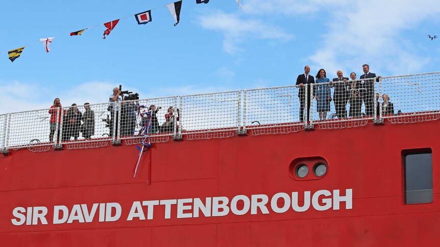 William, Kate and Sir David Attenborough stand on deck of red ship with the latter's name painted the on the side of the ship.