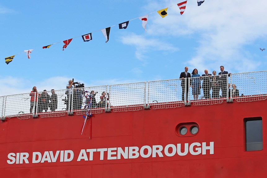 William, Kate and Sir David Attenborough stand on deck of red ship with the latter's name painted the on the side of the ship.