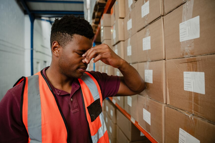 A man in a high-vis shirt looking stressed next to a shelf of boxes.
