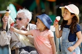 An elderly man eats ice cream on a park bench with two young children.