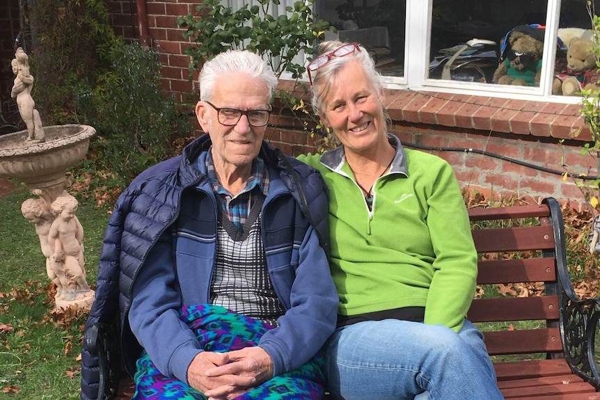 An elderly man and a middle aged woman with smiles on their faces, sitting on a park bench.