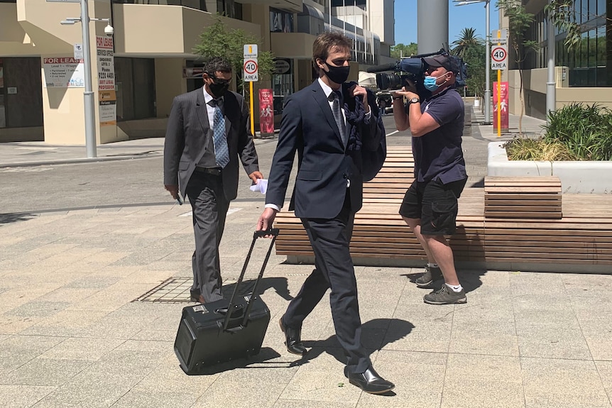 A wide shot of a man wearing a suit and face mask walking outside court in front of another man and a TV camera operator.