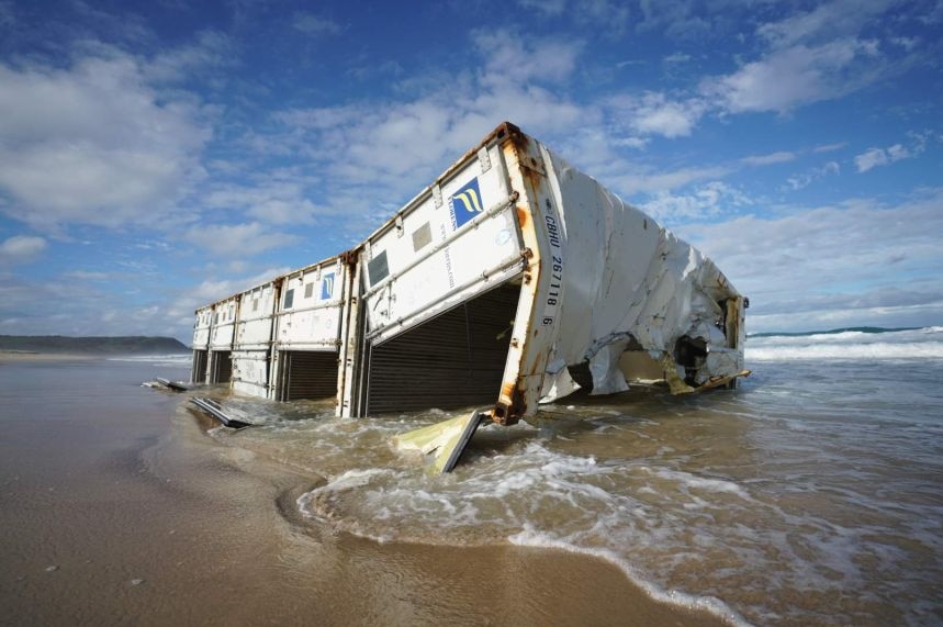A semi-crushed white shipping container sits on the shore at a beach