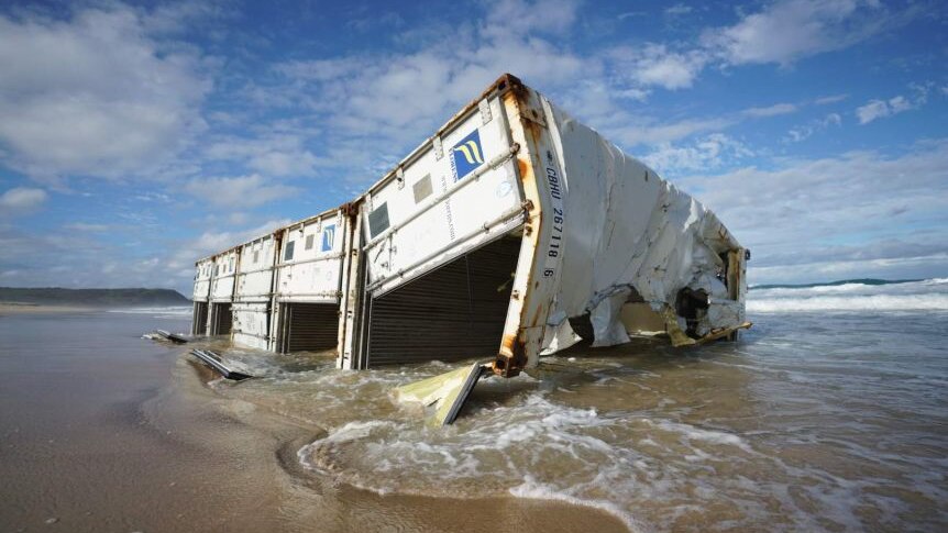 A semi-crushed white shipping container sits on the shore at a beach