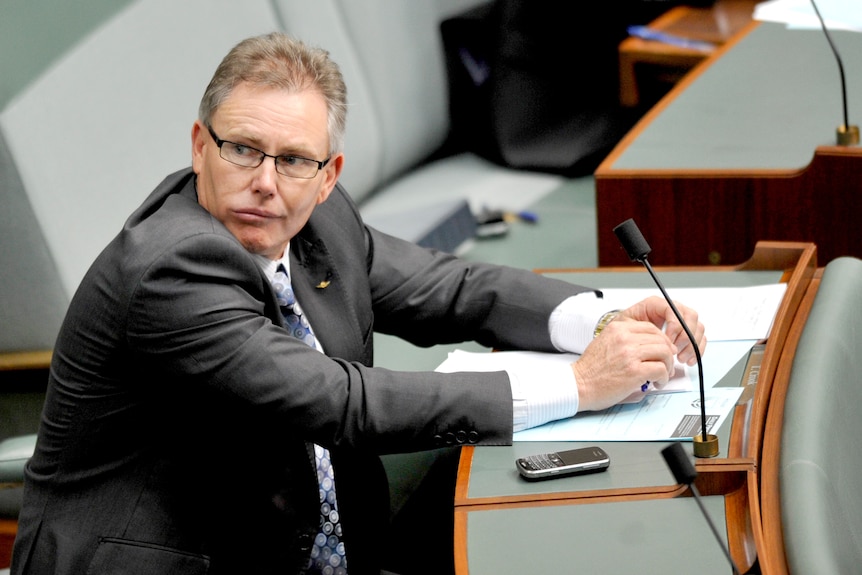 A bespectacled politician in a dark suit sits in parliament.