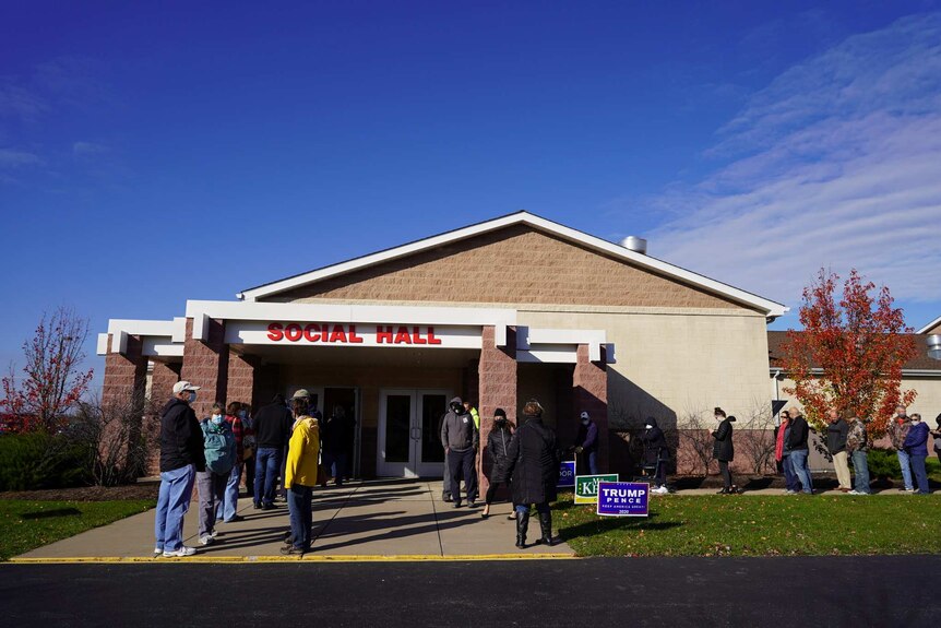 people line up outside a building with political campaign signs on the lawn and a sign reading social hall
