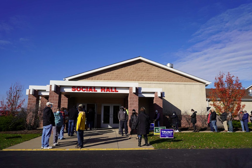 people line up outside a building with political campaign signs on the lawn and a sign reading social hall