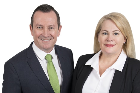 A man in a suit, white shirt and green tie stands with a woman in a black jacket and white shirt next to a sign.