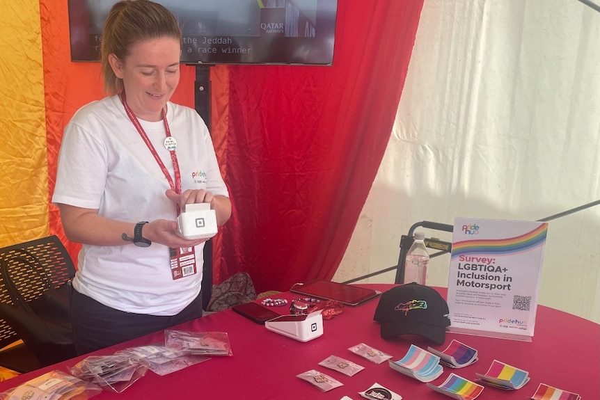 A woman in a white t-shirt is in a marquee selling merchandise on a table in front of her.