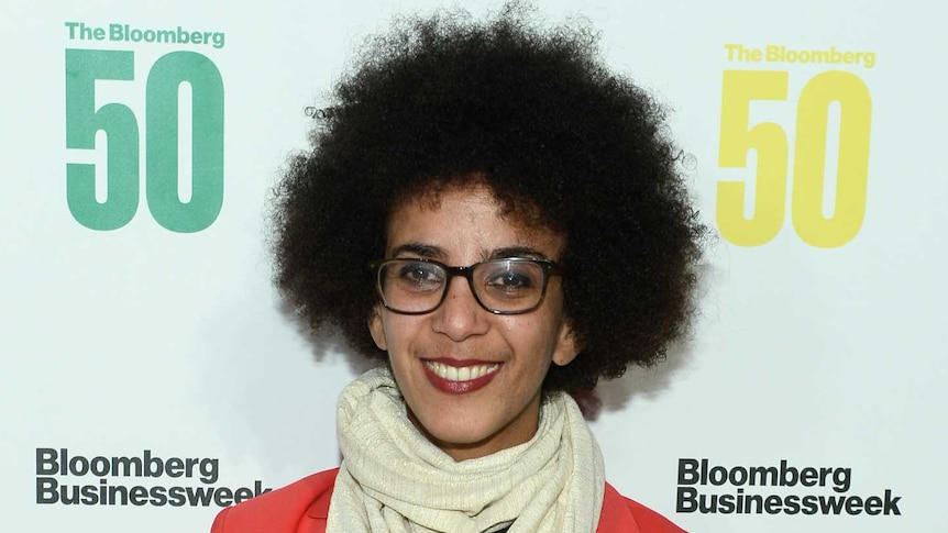You view a woman of African descent with an Afro smiling in a red blazer, standing in front of a media logo wall.
